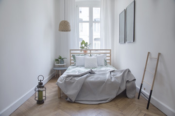 Modern scandinavian sunny bedroom with plants , floral pattern bedding and pilows. Space with white walls and brown wooden parquet. 