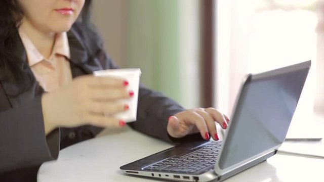 A woman drinks a drink from a disposable cup and works on a laptop. Slow mo. Camera moves