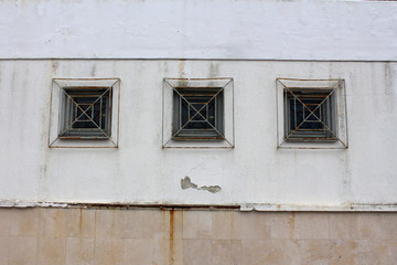 Three wooden frame rectangle glass windows with partially rusted metal bars mounted on dilapidated wall outside