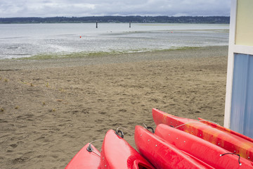 Empty red plastic recreational kayaks for rent or hire, stored on sandy beach after hours on a rainy day. Crescent Beach, Surrey, British Columbia, Canada.