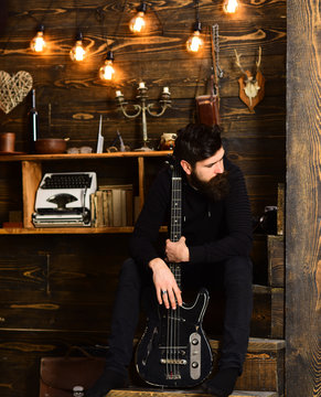 Connection through music. Guy sits thoughtful dreamy in cozy warm atmosphere. Man bearded musician enjoy evening with bass guitar, wooden background. Man with beard holds black electric guitar