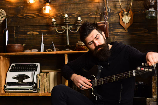 Guitar as hobby. Man bearded musician enjoy evening with bass guitar, wooden background. Guy in cozy warm atmosphere play favourite music. Man with beard holds black electric guitar