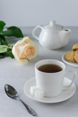 A cup of tea in a white mug, a cookie on a plate, a teapot with tea and a cream rose for a good start to the day