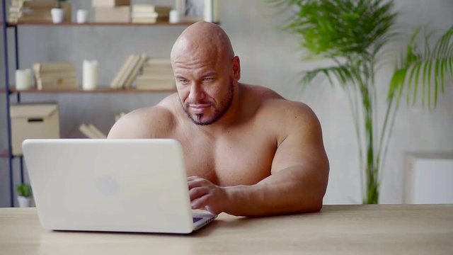 bald man with big muscles sits naked at the table and leads a video link on the laptop to train the body constructs