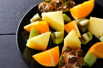 Fruit salad on a black plate on wooden boards, pineapples, oranges and kiwi on a dark background, salad of tropical fruits for breakfast, vegetarian food, copy space