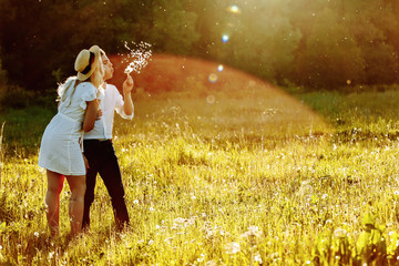Couple walking in summer field. Young man and woman blowing dandelions at sunset.