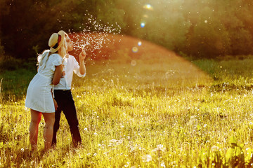 Plakat Couple walking in summer field. Young man and woman blowing dandelions at sunset.