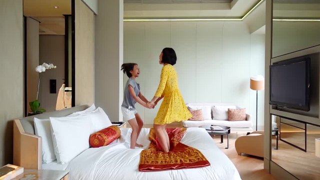 Slow motion of happy woman and her daughter jumping on the bed while holding hands together in the hotel room