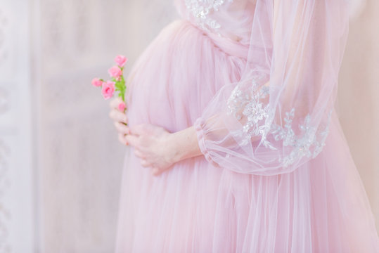 Beautiful pregnant woman in rich pink dress holds hands on her belly posing in luxury white room