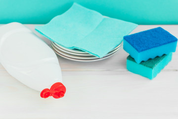 Bottle of dish washing, sponges, utensils on white wooden table against a green wall background,