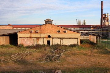 Fototapeta na wymiar Broken down old industrial brick building with wooden roof, metal air vents, high uncut grass around it, old metal chimneys and building in background