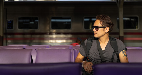 a traveller, backpacker man in casual clothes and sunglasses with a camera, sitting and waiting for a train at platform train station