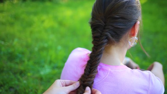 Closeup view of young girl sitting on red picnic blanket outside with her back to camera and female hands of her friend helping her with making braid. 
