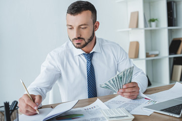 handsome business adviser counting money in office and writing something to notebook