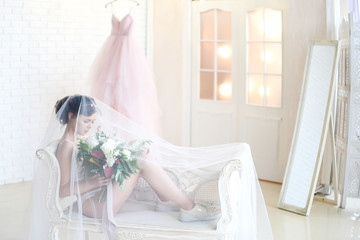 Bride in white lingerie and keds sits on a white couch under the veil