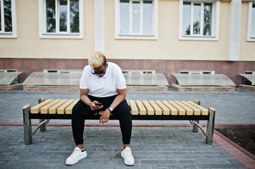 Stylish arabian muslim boy with originally hair and sunglasses posed on streets, sitting on bench and listening music from earphones of phone.