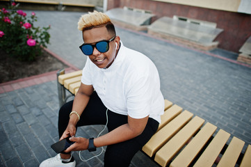 Stylish arabian muslim boy with originally hair and sunglasses posed on streets, sitting on bench and listening music from earphones of phone.