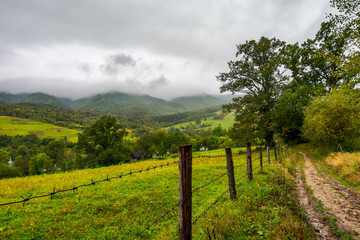 Fototapeta na wymiar road along the fence in rural outskirts. pasture behind the barbwire. mountainous countryside on a dull day with overcast sky. village down in the valley. some old oak trees along the path
