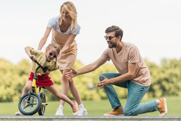 happy parents looking at cute little daughter riding bicycle in park