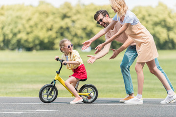 happy parents teaching little daughter riding bicycle in park