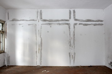 Large moisture patch on white wall of abandoned building in shape of two crosses with dilapidated windows and floor covered in broken glass and dirt