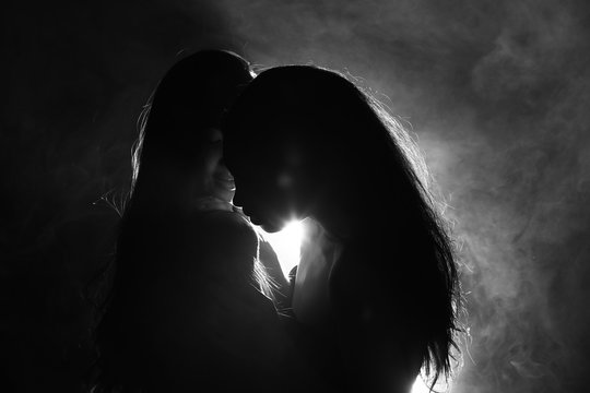Silhouette of two sexy woman kissing holding in darkness through light and smoke