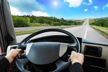 Truck dashboard with driver's hands on the steering wheel on the countryside road against sky with sun