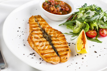 The large salmon steak red fish on the grill with lemon, sauce with vegetables and greens on white plate