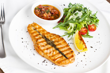The large salmon steak red fish on the grill with lemon, sauce with vegetables and greens on white plate