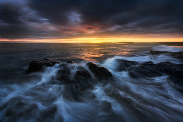 Long exposure of a dramatic and dark, coastal sunset with rocks low in the foreground. The sunlight is glowing across the horizon. The clouds are dark blue/purple and hanging low.