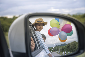 A young child smiles from a car window holding colorful balloons, showcasing joy during a travel adventure. The mirror reflection adds depth to the feeling of happiness and freedom experienced during 