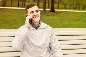 Молодой парень европейской внешности сидит на лавочке говорит по телефону и смеется