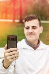 Молодой парень сидит на белой  лавочке, улыбается и делает селфи