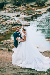 Romantic outdoor wedding shot of the fashionable newlywed couple hugging at the river bank. Bride and groom kiss and hug each other behind beautiful landscape. After wedding ceremony.