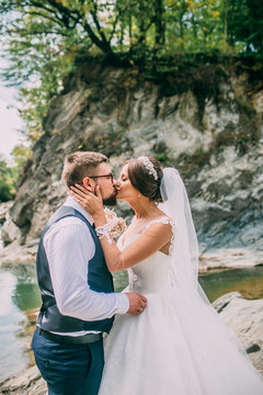 Romantic outdoor wedding shot of the fashionable newlywed couple hugging at the river bank. Bride and groom kiss and hug each other behind beautiful landscape. After wedding ceremony.