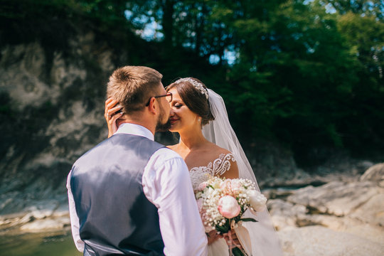 Romantic outdoor wedding shot of the fashionable newlywed couple hugging at the river bank. Bride and groom kiss and hug each other behind beautiful landscape. After wedding ceremony.