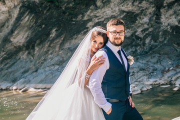 Romantic wedding moment of newlywed couple hugging near blue mountain river, sensual groom embracing gorgeous bride from behind river. Wedding day for man and woman.