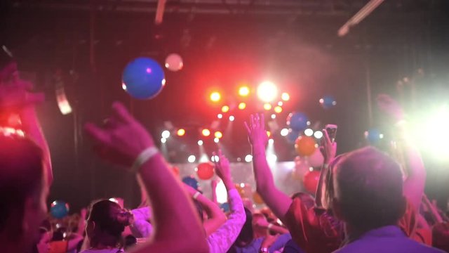 Crowd of people dancing raising hands at a concert - multi-colored balloons flying around the concert hall