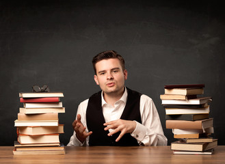 A young teacher in glasses sitting at classroom desk with pile of books in front of clean blackboard back to school concept.