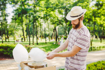 Fototapeta premium photo theme small business cooking sweets. A young man with a beard of a Caucasian trader in the hat the owner of the outlet makes candy floss, fairy floss or Cotton candy in the summer park