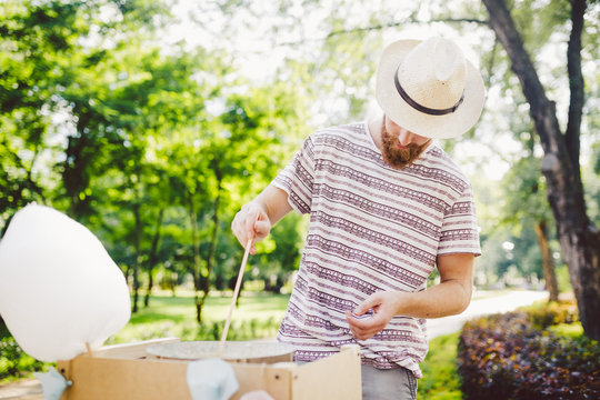 Photo Theme Small Business Cooking Sweets. A Young Man With A Beard Of A Caucasian Trader In The Hat The Owner Of The Outlet Makes Candy Floss, Fairy Floss Or Cotton Candy In The Summer Park