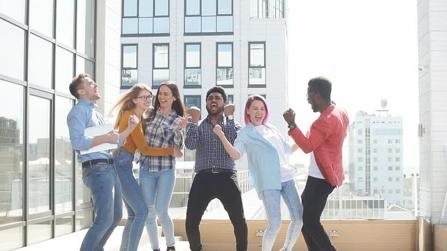 Multiracial heterosexual group of students having fun on roof party dancing and listening music at daytime on the roof area against glass office buildings background