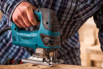 Close up of an experienced carpenter in work clothes and small buiness owner  is carving a wooden board on an  modern  hand drill in a workshop , in the background a lot of tools