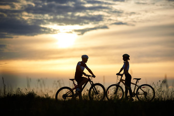 Two cyclists, guy and girl standing opposite each other and posing with bikes. Silhouettes of couple, looking at each other, against amazing and wonderful background of sky with clouds and sunset.