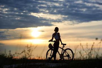 Obraz na płótnie Canvas Silhouette of sporty cyclist, wearing sportswear and standing near her bicycle on trail. View of incognito woman enjoying nature and observing wonderful landscapes and amazing sunset. Back view