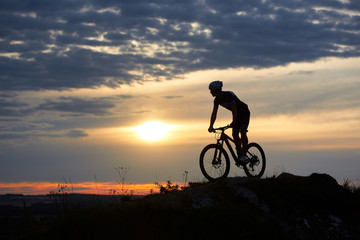 Fototapeta na wymiar Silhouette of male cyclist wearing a helmet and sports clothes riding a mountain bike on rocks in the mountains against a beautiful sunset with a bright sun and sky in the clouds.