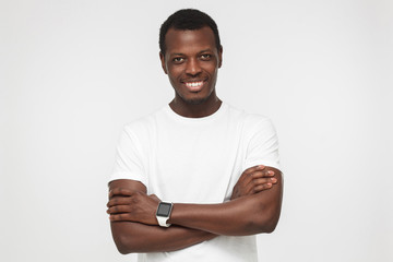 Portrait of smiling handsome afican american man in blank white t shirt and smart watches, standing with crossed arms isolated on grey background