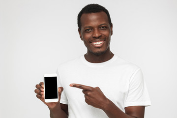 Happy african american man wearing white t shirt isolated on grey background demonstrating blank smartphone and pointing to screen. Copyspace for advertising products and services.
