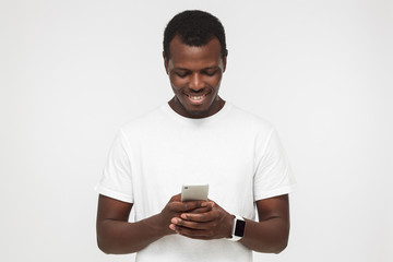Closeup photo of African American man standing isolated on gray background looking at screen of cellphone, browsing web pages and smiling nicely while chatting