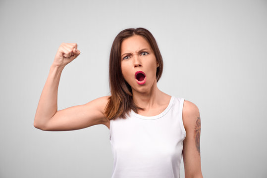 Horizontal shot of casually dressed young lady shouting fiercely and tensing bicep, showing strength and power. Pretty girl with fit body demonstrating muscles. Fitness, energy, readiness concept